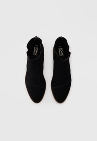 O pereche de bocanci negri din suede TOMS, cu vârfuri ascuțite și tălpi maro, văzuți de sus pe un fundal alb simplu.