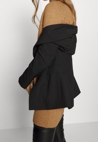 Černá bunda s prostornou kapucí, nošená přes přiléhavé, světle hnědé pletené šaty. Šaty mají žebrovanou strukturu; oblečení doplňují černé vysoké kozačky.