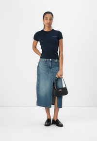 T-shirt di cotone navy, gonna midi in denim con spacco frontale e borsa nera strutturata. Modella indossa mocassini neri, capelli intrecciati.