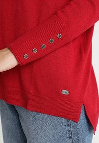 Maglione rosso a maglia con polsini a costine, dotato di cinque bottoni grigi. L'orlo è leggermente più lungo ai lati, con una piccola etichetta del marchio in basso.