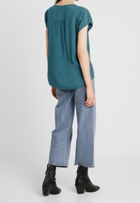 Haut à manches courtes teal avec des plis au dos, associé à un jean court bleu clair et des bottines noires à zips. Texture de tissu lisse.