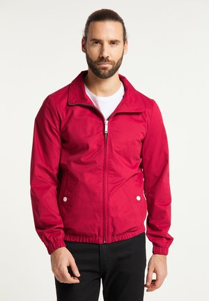 Mann mit Bart und zurückgebundenem Haar, der eine rote Jacke mit Reißverschluss über einem weißen Hemd und schwarzen Hosen trägt, vor einem einfachen hellen Hintergrund.