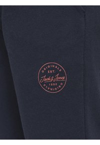 Námorně modrá bavlněná látka s kulatým červeným logem, na kterém je vytištěno "Jack & Jones" a "Založeno 1990". Hladká textura, neformální design.