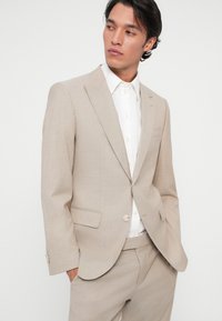 Beige, szabott öltöny finom textúrával, egysoros zakó, kerekített hajtókák, és fehér ing. Nincsenek látható minták vagy díszítések.