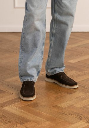 Persoon draagt lichtblauwe spijkerbroek en donkerbruine suède instapschoenen, staande op een houten parketvloer.