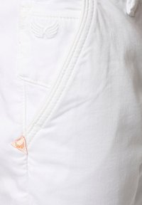 Primer plano de una tela blanca con una textura sutil, que presenta un bolsillo en el lado derecho y una pequeña etiqueta triangular naranja con un logotipo de ala en relieve.