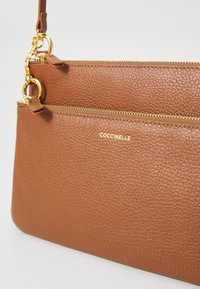 Braune, strukturierte Leder-Clutch mit goldfarbenen Beschlägen, ausgestattet mit zwei Reißverschlussfächern und dem eingeprägten Markennamen "Coccinelle".