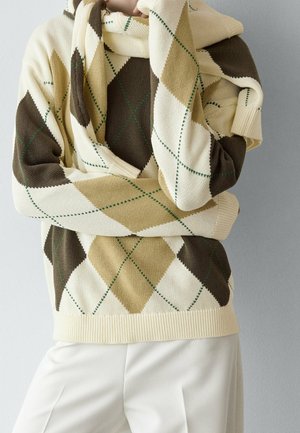 Personne portant un pull à motif losanges argyle crème, beige et marron avec une écharpe assortie et un pantalon blanc sur un fond gris uni.