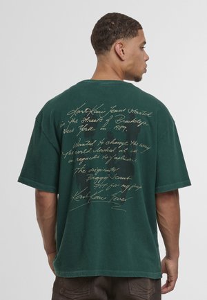 T-shirt en coton vert surdimensionné avec un grand texte manuscrit contrastant au dos, coupe décontractée et épaules tombantes.