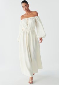 Una donna cammina in un abito bianco fluente con spalle scoperte e vita stretta, accessoriate con orecchini, su uno sfondo chiaro.