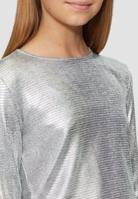 Silberner metallischer Oberteil mit einem strukturierten Gittermuster, ausgestattet mit einem Rundhalsausschnitt und langen Ärmeln, gefertigt aus einem glänzenden, reflektierenden Material.