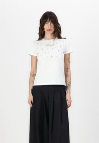 Camiseta blanca de manga corta con adornos plateados decorativos y escote redondeado, combinada con pantalones negros de cintura alta.