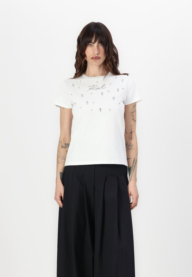 Camiseta blanca de manga corta con adornos plateados decorativos y escote redondeado, combinada con pantalones negros de cintura alta.