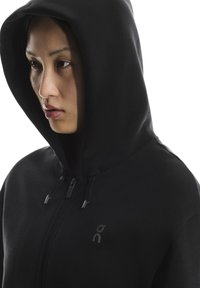 Hanorac cu fermoar negru, cu glugă proeminentă, logo subtil pe piept și material texturat. Detaliile cu fermoar în față sporesc funcționalitatea.