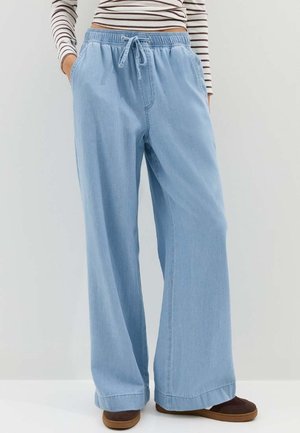 Personne portant un pantalon en denim bleu clair à jambes larges avec taille à cordon, un haut à manches longues rayé, et des chaussures marron à enfiler, debout les mains dans les poches.