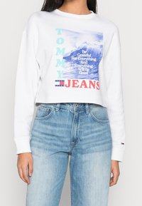 Vit kortärmad sweatshirt med blå och röd grafisk tryck, som har texten "TOMMY" och "JEANS", tillsammans med vågmotiv. Bärs med ljusblå jeans.