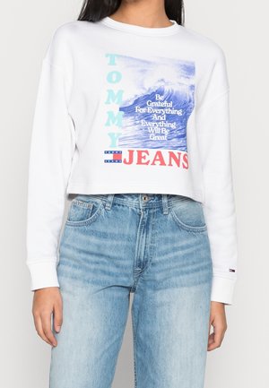 Vit kortärmad sweatshirt med blå och röd grafisk tryck, som har texten "TOMMY" och "JEANS", tillsammans med vågmotiv. Bärs med ljusblå jeans.