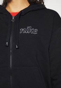 Felpa nera con zip realizzata in morbido tessuto, con un logo "nike" ricamato bianco sul petto e una tasca frontale, dotata di cordini regolabili.