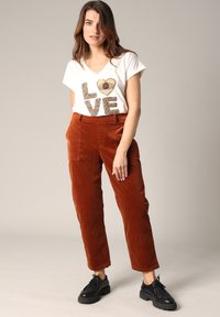 T-shirt blanc à col en V avec un graphique "LOVE" en lettres dorées et brunes, associé à un pantalon en velours côtelé couleur rouille et des chaussures noires à lacets.