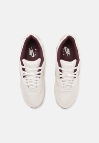 Baltās Nike kedas ar teksturētu sietu augšdaļu, bordo oderei un baltiem šņorēm. Uz mēles ir Nike Air zīmols.