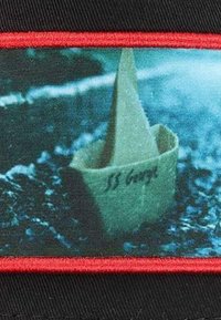 Beigefarbener Papierboot mit der Aufschrift "SS George" auf der Seite, schwebt auf einer dunklen, strukturierten Oberfläche, umrahmt von rotem Nähen auf schwarzem Hintergrund.