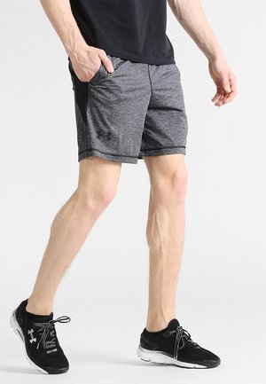 Mężczyzna ubrany w szare sportowe szorty Under Armour i czarne buty do biegania Under Armour, stoi z jedną ręką w kieszeni.