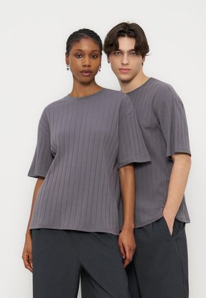 Δύο μοντέλα στέκονται κοντά, φορώντας ταιριαστά χαλαρά γκρι μπλουζάκια με κοντά μανίκια και ριβ ύφανση και σκούρα παντελόνια, σε απλό λευκό φόντο.