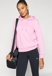 Pink hooded sweatshirt with drawstrings and front logo, paired with black leggings featuring white branding, held with a black bag.