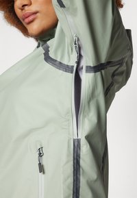 Veste imperméable vert clair avec des accents foncés contrastants, des poches zippées et un col haut. Le matériau semble lisse et léger.