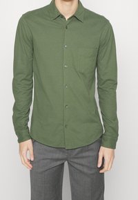 Zelená košile s dlouhým rukávem a knoflíky vyrobená z měkké tkaniny, obsahující kapsu na prsou, špičatý límec a tmavé knoflíky.