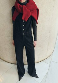 Mono de mezclilla negro con piernas anchas, botones plateados y bolsillos delanteros, complementado con una gran bufanda de punto acanalada roja.