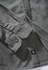 Grå skjorta med en slät textur, knäppta manschetter, kontrasterande knappar och en etikett med en stiliserad 'B' på sidoverkan.