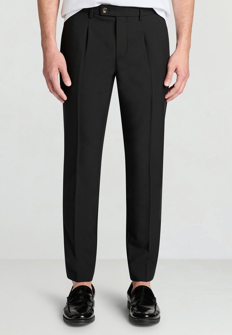 Pantaloni neri sartoriali con taglio dritto, dotati di piega frontale e chiusura con bottone, abbinati a mocassini neri lucidi.