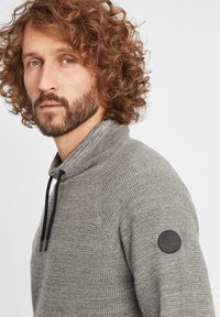 Szary sweter z dzianiny z halowym zamkiem, z teksturowanym materiałem, czarnymi sznurkami do regulacji i okrągłą czarną naszywką z logo na ramieniu.