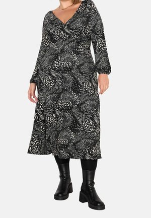 Nainen pukeutunut mustavalkoiseen kuvioituun pitkähihainen midi-mekkoon, mustiin legginsseihin ja mustiin paksupohjaisiin saappaisiin.