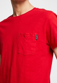 Rød bomulds T-shirt med rund hals og korte ærmer. Har en brystlomme og et lille mærke på lommen. Lidt tekstur er synlig.