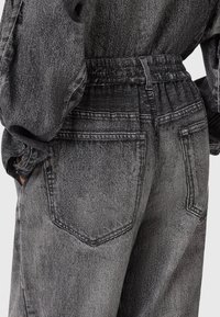 Pantalones de mezclilla negra con lavado ácido, con cintura elástica y bolsillos traseros visibles, llevados con una camisa de mezclilla a juego con las mangas arremangadas.