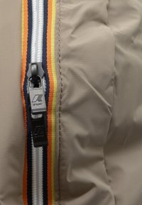 Giacca beige con zip e superficie testurizzata, caratterizzata da una zip con hardware nero e strisce colorate arancioni, blu navy e gialle.