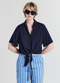 Chemise marine à manches courtes avec un détail de nœud à l'avant et une poche ; associée à un pantalon bleu clair à rayures avec une taille à cordon. Lunettes de soleil foncées portées.