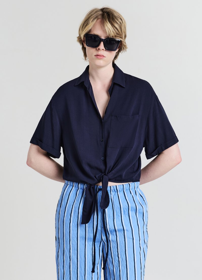 Chemise marine à manches courtes avec un détail de nœud à l'avant et une poche ; associée à un pantalon bleu clair à rayures avec une taille à cordon. Lunettes de soleil foncées portées.