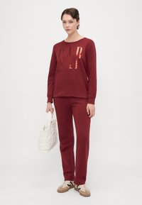 Burgundská mikina s reflexním logem "GAP", odpovídající kalhoty a béžové tenisky. Drží bílou texturovanou tašku. Minimalistický design.