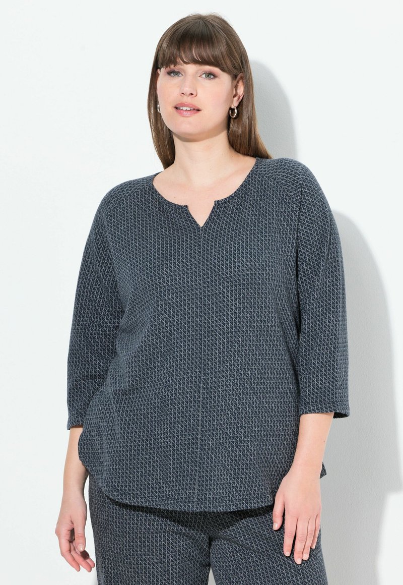 Una donna con capelli castani lisci indossa un top a maniche lunghe grigio scuro con motivo e pantaloni coordinati, in piedi contro uno sfondo bianco.