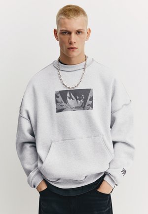 Felpa grigia con tasca frontale e grafica stampata sul petto. Il design include una collana a catena e jeans neri.