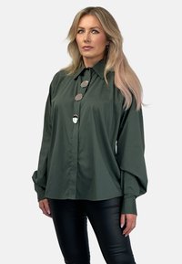 Ciemnozielona koszula z guzikami i kołnierzykiem, z trzema dużymi srebrnymi guzikami i luźnymi rękawami. Noszona z czarnymi skórzanymi spodniami.