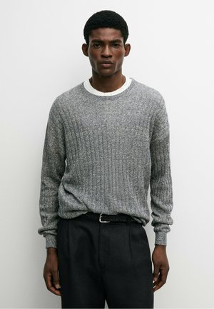 Jeune homme aux cheveux courts et bouclés portant un pull côtelé gris sur une chemise blanche et un pantalon noir, sur un fond blanc uni.