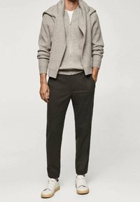 Grijze gebreide jas met rits over een grijze geribbelde trui, gecombineerd met donkere broek en witte sneakers. Soepele textuur en casual pasvorm.