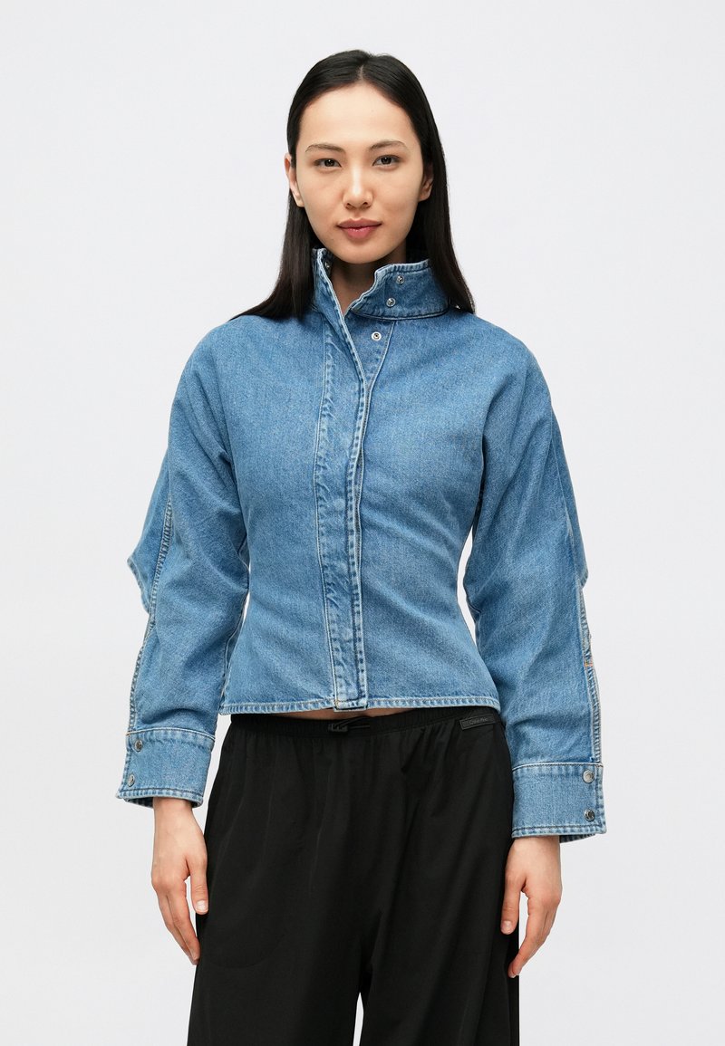 Gaiši zils džinsa jaka ar augstu apkakli, priekšējo pogu aizdari un brīvu piegriezumu. Raksturīgas sānu šķēlumi un izšūtas dekoratīvas detaļas.