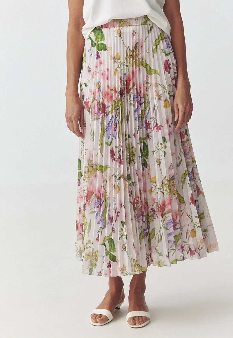 Femme portant une jupe plissée longueur cheville avec imprimé floral multicolore, un haut blanc et des sandales blanches à bouts ouverts.