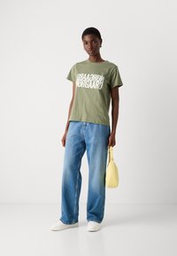 Olivgrünes Baumwoll-T-Shirt mit weißem grafischem Text, kombiniert mit lässigen blauen Jeans und einer kleinen gelben Tasche.