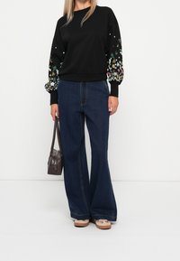 Femme portant un pull noir avec des manches à motif floral, un jean large bleu foncé, des baskets beiges, et tenant un sac à main marron foncé.